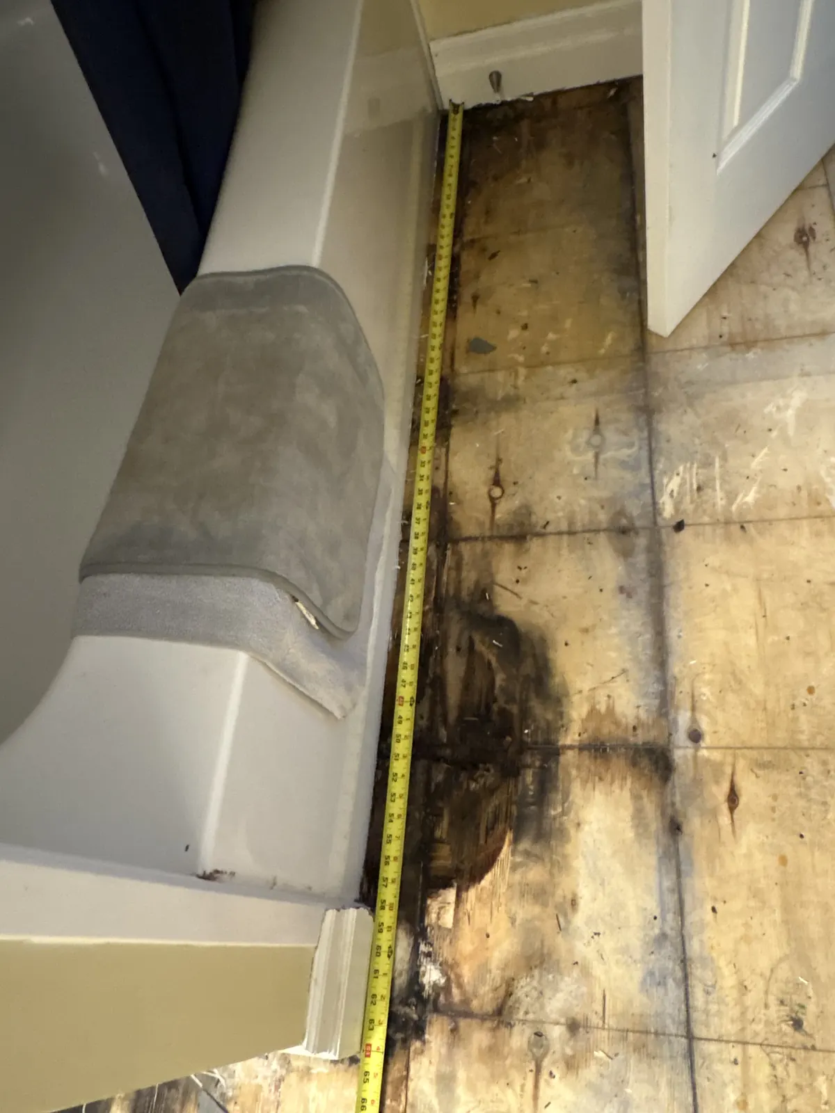Water-damaged plywood subfloor exposed beside a bathroom vanity, tape measure laid across it